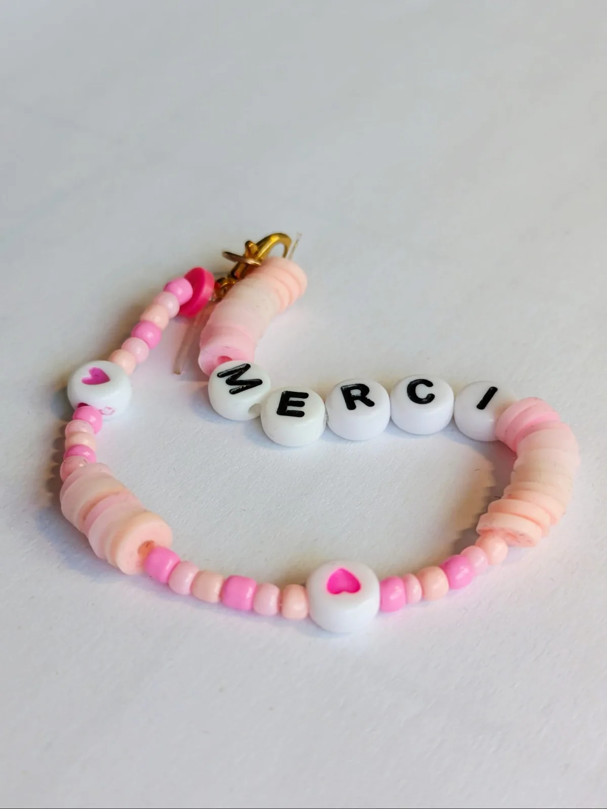 Bracelet artisanal en perles roses et pêche formant le mot MERCI avec une breloque cœur rose, fermoir mousqueton doré